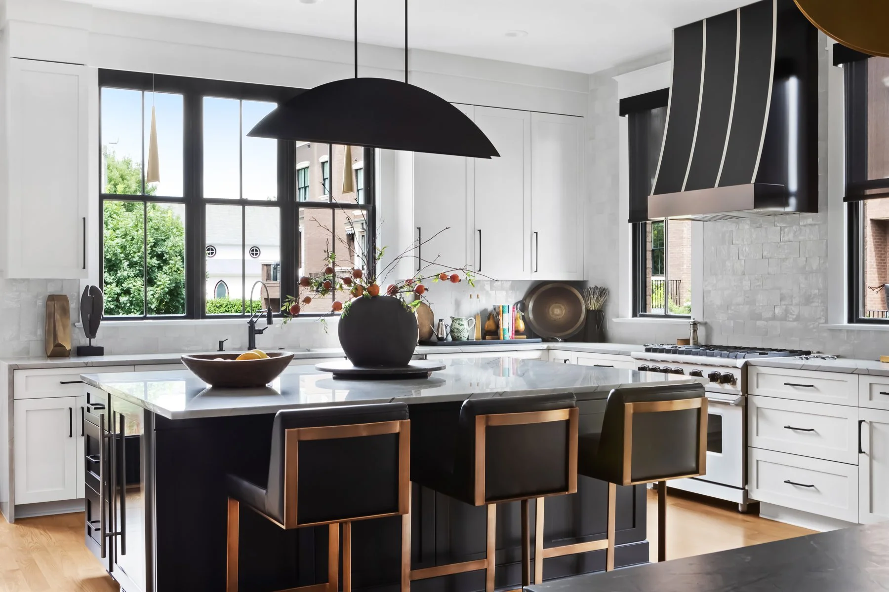 Townhome kitchen with white cabinetry, a marble waterfall island, black tapered range hood and brass-framed leather barstools
