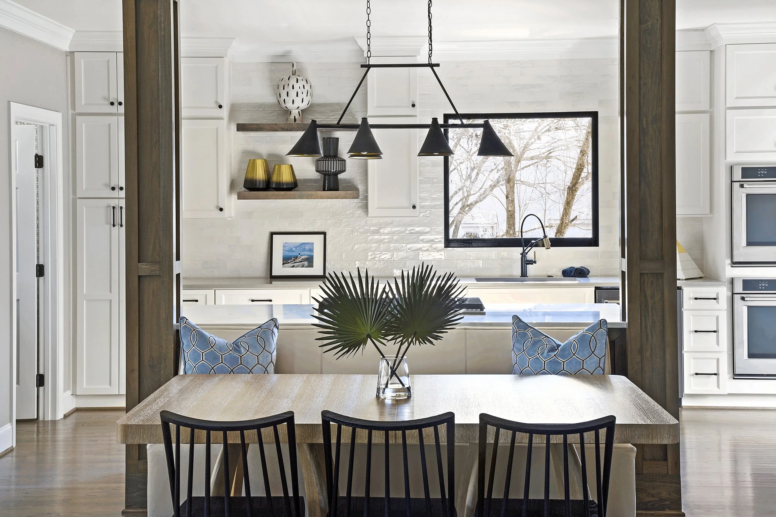 White kitchen framed by timber posts with a black cone-shade linear pendant, banquette seating, oak table and black spindle chairs